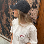 Person wearing a black cap and white t-shirt with a graphic design, standing against a textured wall.