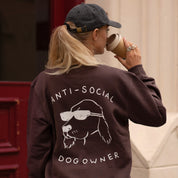 Person wearing a sweatshirt with 'ANTI-SOCIAL DOG OWNER' text and dog graphic, holding a coffee cup.
