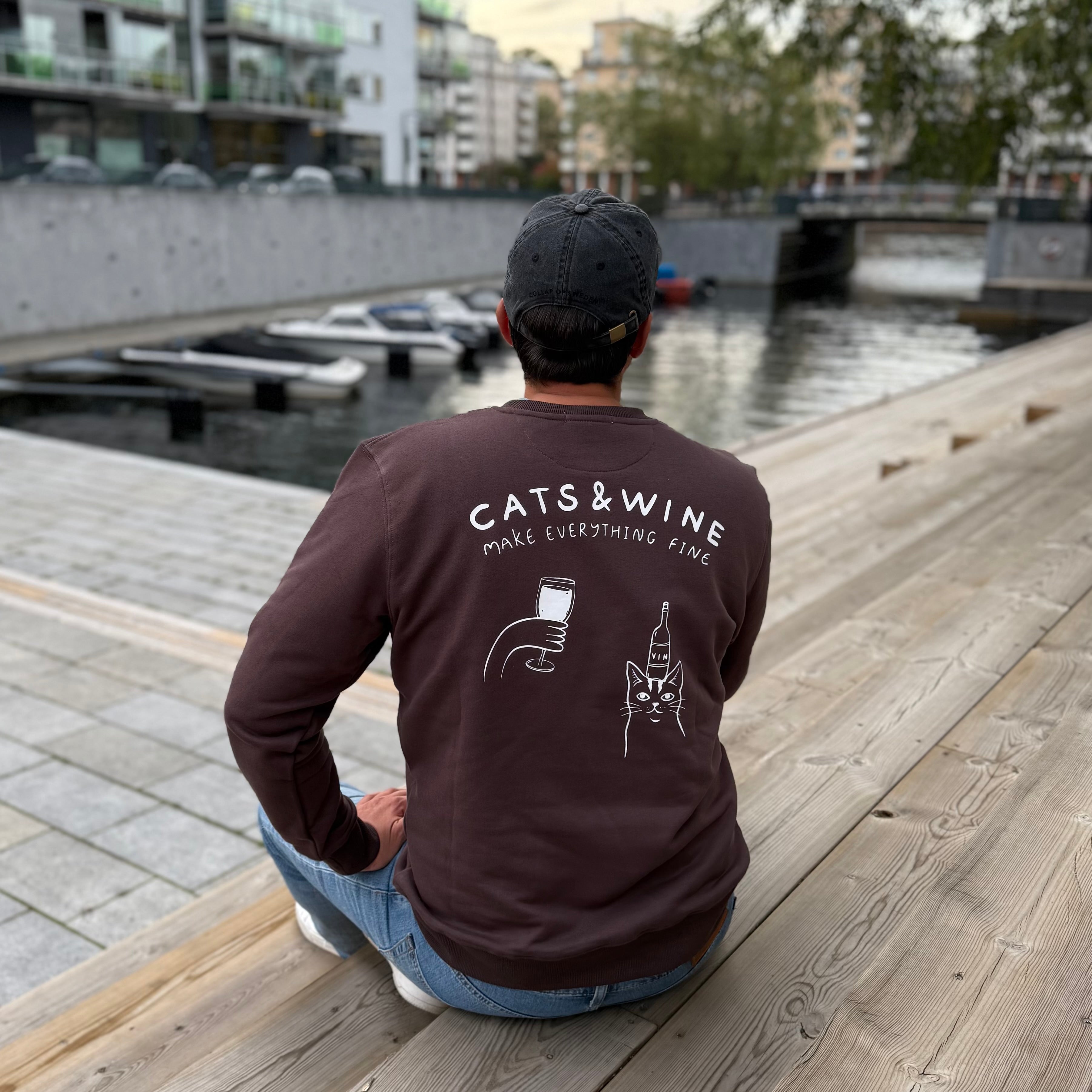 Person wearing a brown long-sleeve shirt with 'Cats & Wine Make Everything Fine' text and graphics, sitting on a wooden dock by a waterfront.